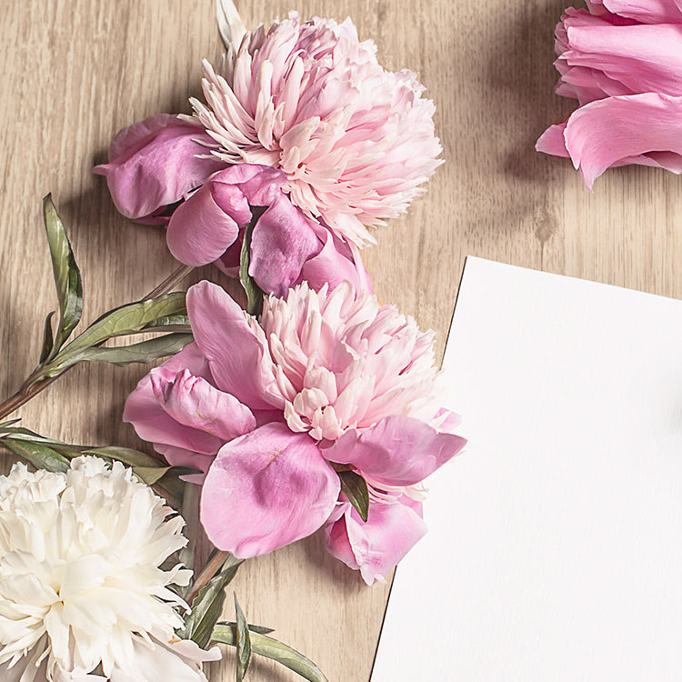 Pink and white peonies rest on a wooden table beside a blank white piece of paper, with green leaves scattered around, creating a serene and elegant arrangement.