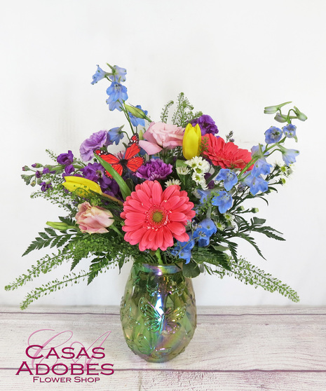 Iridescent vase with butterflies on it with pink gerbera daisy, blue delphinium, pink lisianthus, purple carnations and purple stock